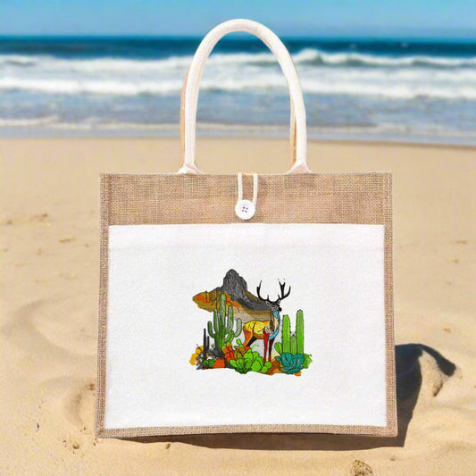 A jute tote bag featuring a colorful embroidered design of a deer in a desert landscape with cacti and mountains.
