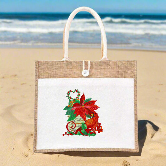 Jute tote bag with white front panel on a sandy beach, featuring embroidered poinsettia, red cardinal, holly, and a small heart wreath.