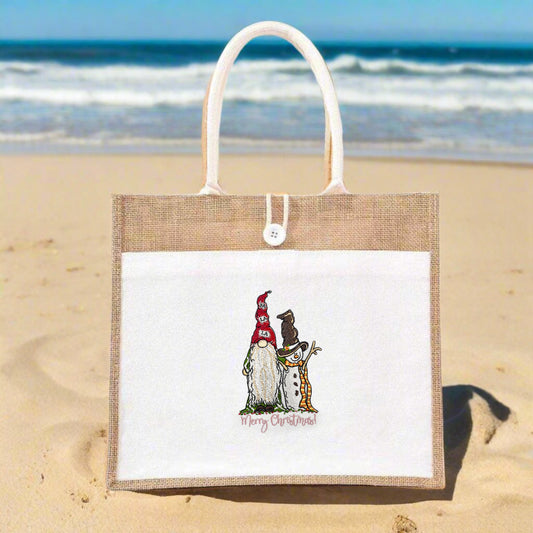 Jute tote bag with white front panel on a sandy beach, featuring an embroidered gnome and snowman above the text “Merry Christmas!”.