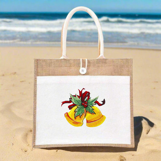 Jute tote bag with white front panel on a sandy beach, featuring embroidered golden Christmas bells with green holly leaves and a red ribbon.