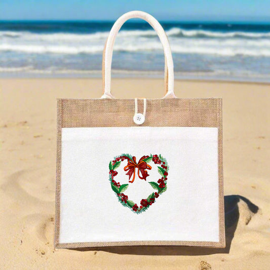 Jute tote bag with white front panel on a sandy beach, featuring an embroidered heart-shaped holly wreath with green leaves, red berries, and a red bow.