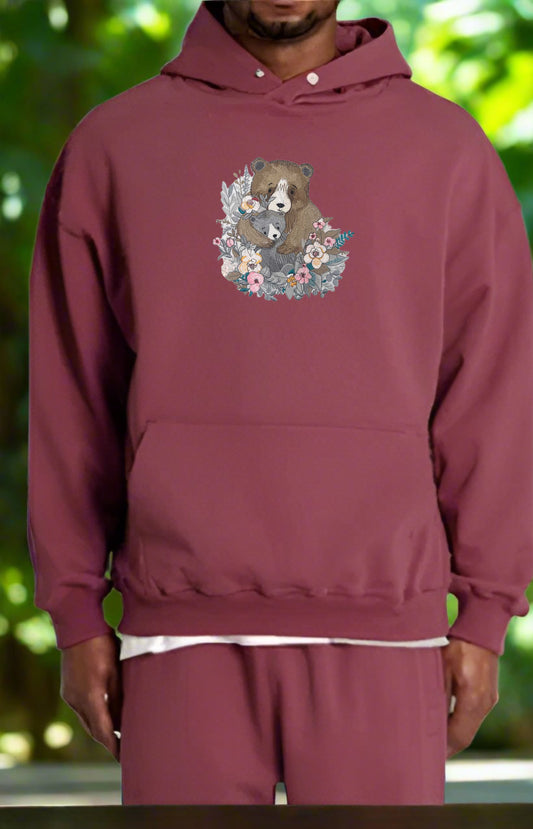 Maroon hoodie with floral bear design worn by a person against a blurred green background