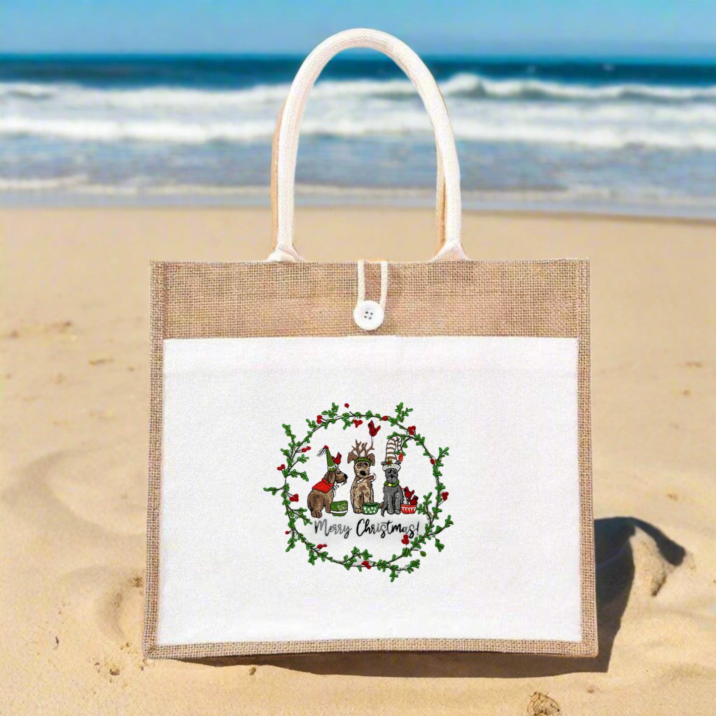 Jute tote bag with white front panel on a sandy beach, showcasing an embroidered wreath of holly with three festive dogs and “Merry Christmas!” caption.
