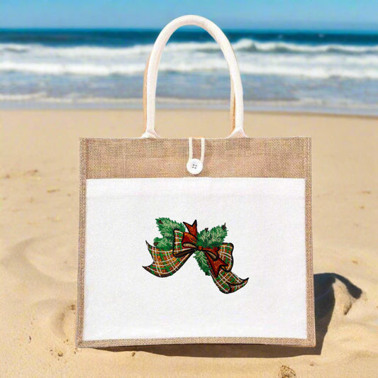 Jute tote bag with white front panel on a sandy beach, displaying an embroidered tartan bow accented with green pine foliage.
