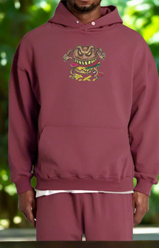 Maroon hoodie with a Cursed Burger Embroidery – Quirky Monster design worn by a person against a blurred green background