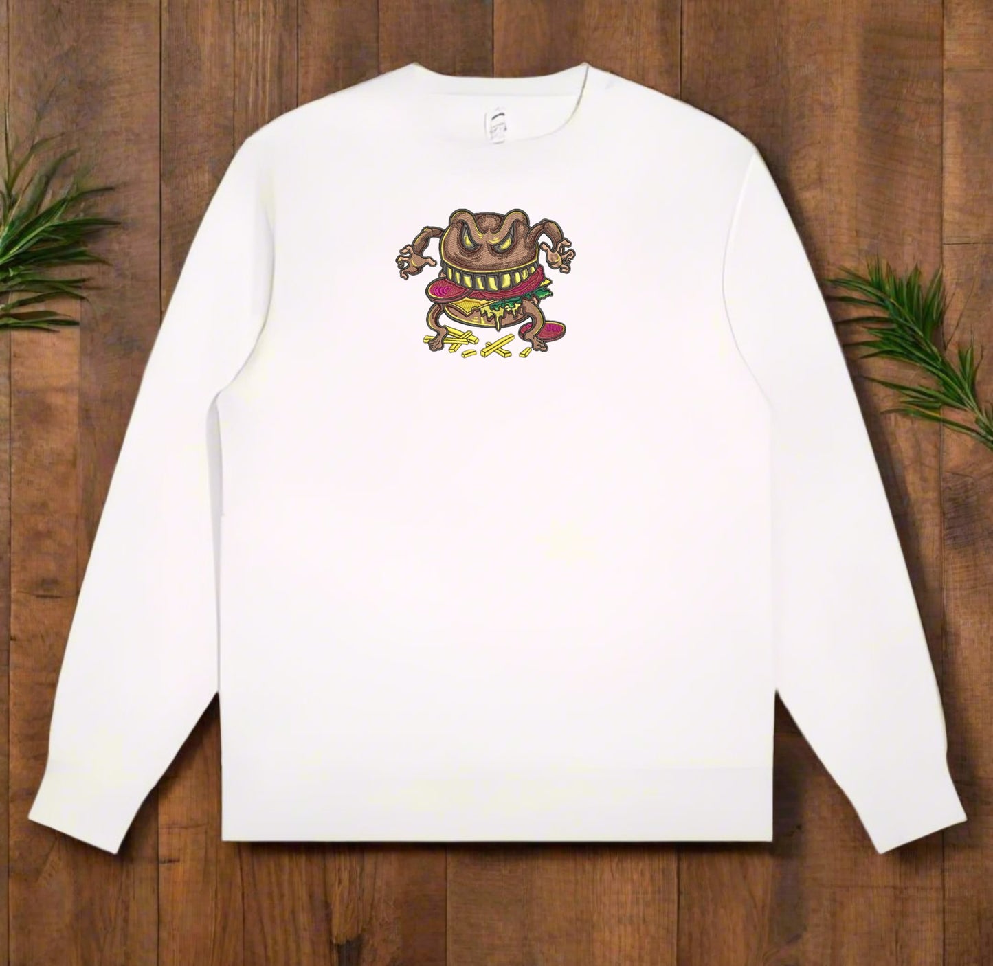 White long-sleeve shirt with a Cursed Burger Monster Embroidery – Funky Foodie design on a wooden surface