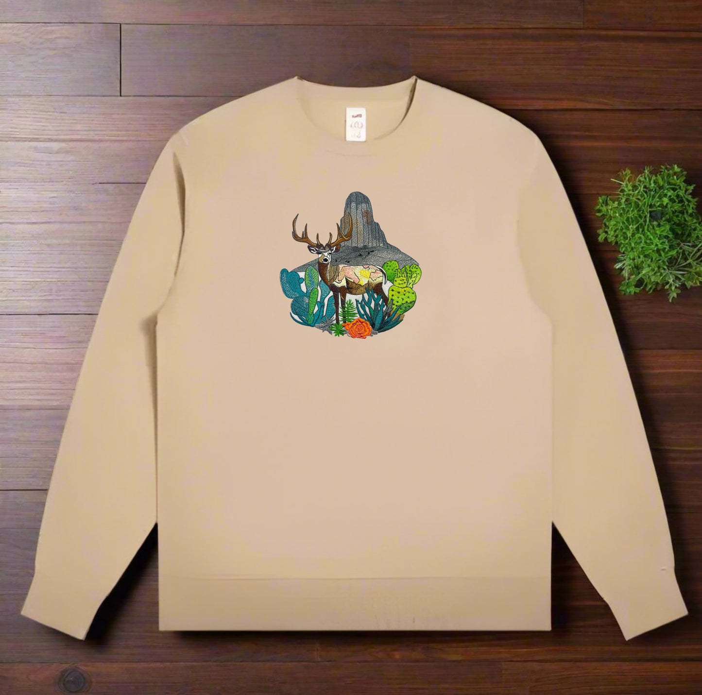 Embroidered crewneck sweatshirt with a design of a mountain deer surrounded by wildflowers and alpine pines against a wooden background.