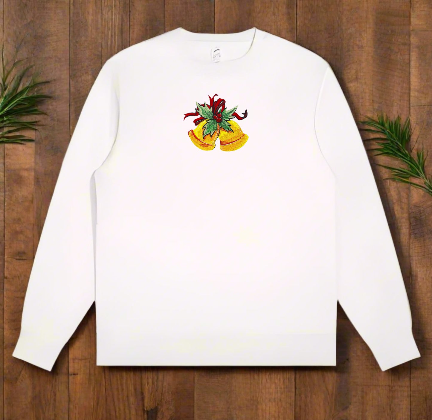 White crewneck sweatshirt on wood background, showcasing embroidered golden holiday bells adorned with holly leaves and a red ribbon