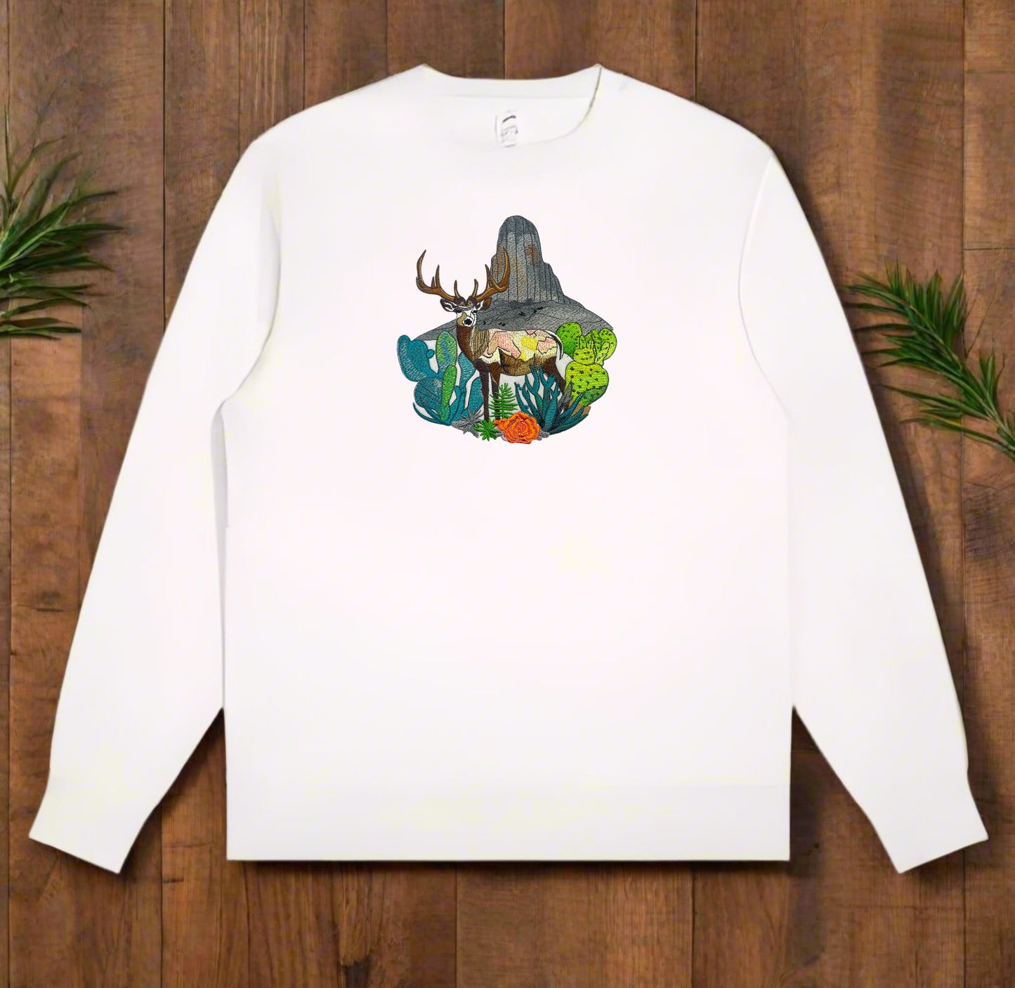 Embroidered crewneck sweatshirt with a design of a mountain deer surrounded by wildflowers and alpine pines against a with wooden background.