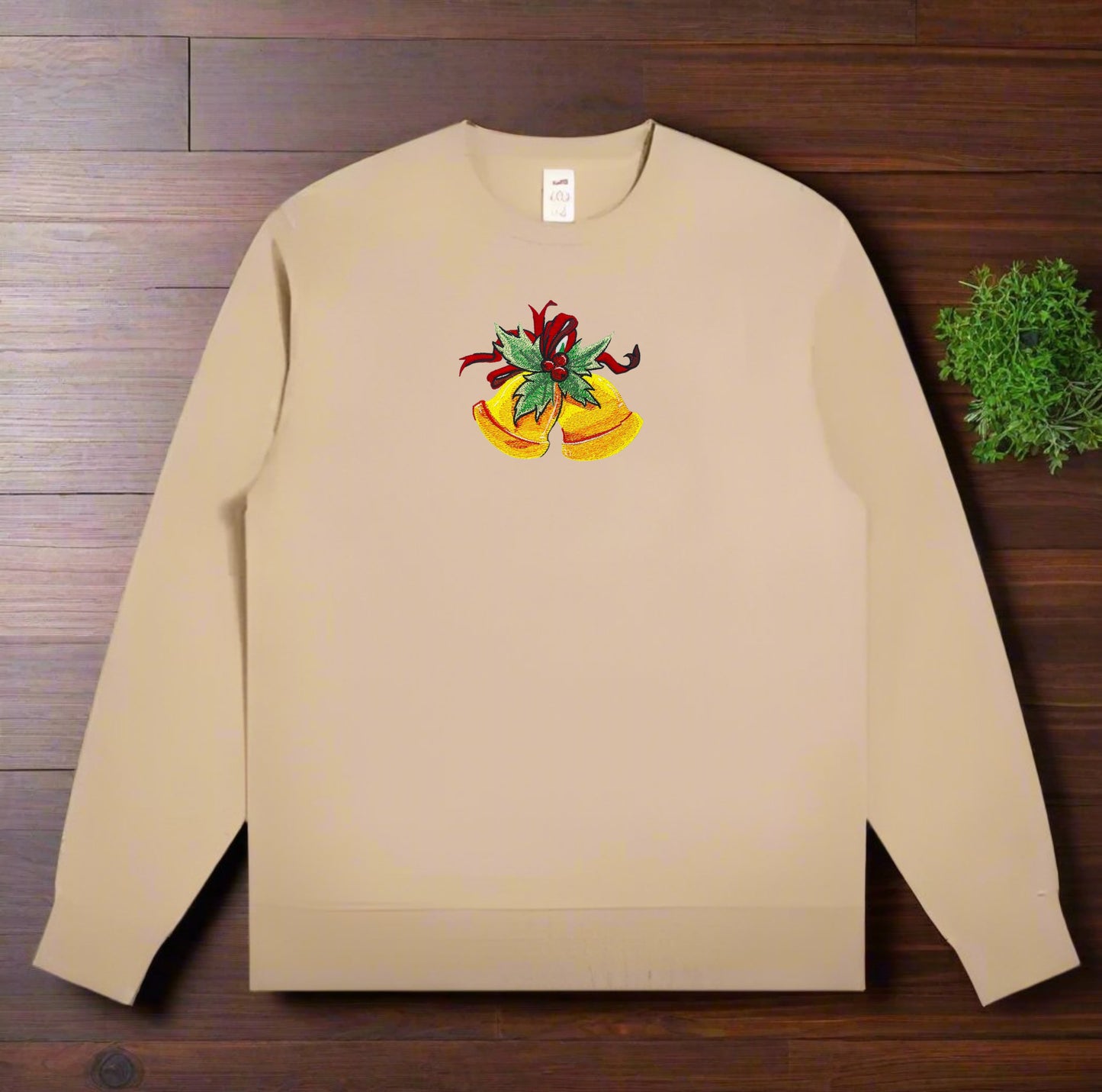 Tan crewneck sweatshirt laid on a wooden surface, featuring embroidered golden Christmas bells with green holly leaves and a red ribbon centered on the chest.