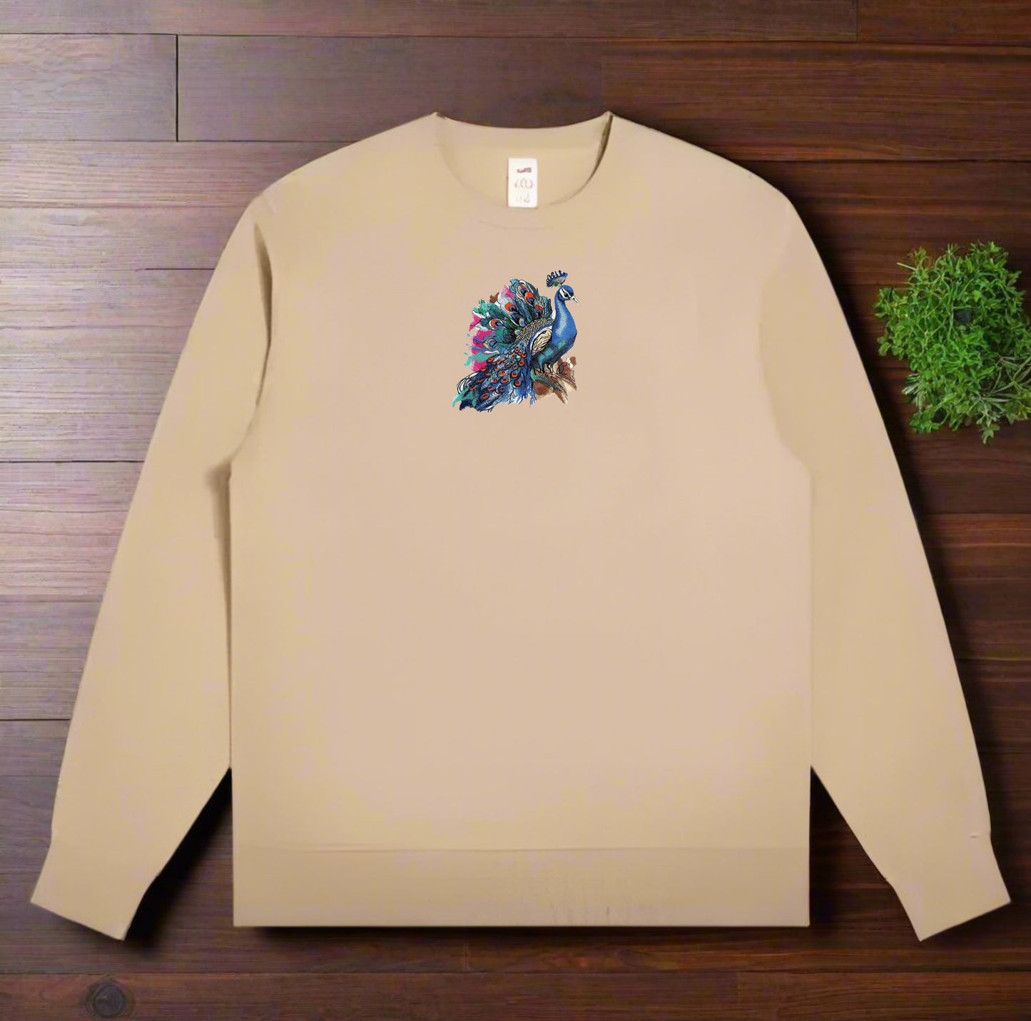 Beige long-sleeve sweatshirt with a colorful peacock design on a wooden surface.