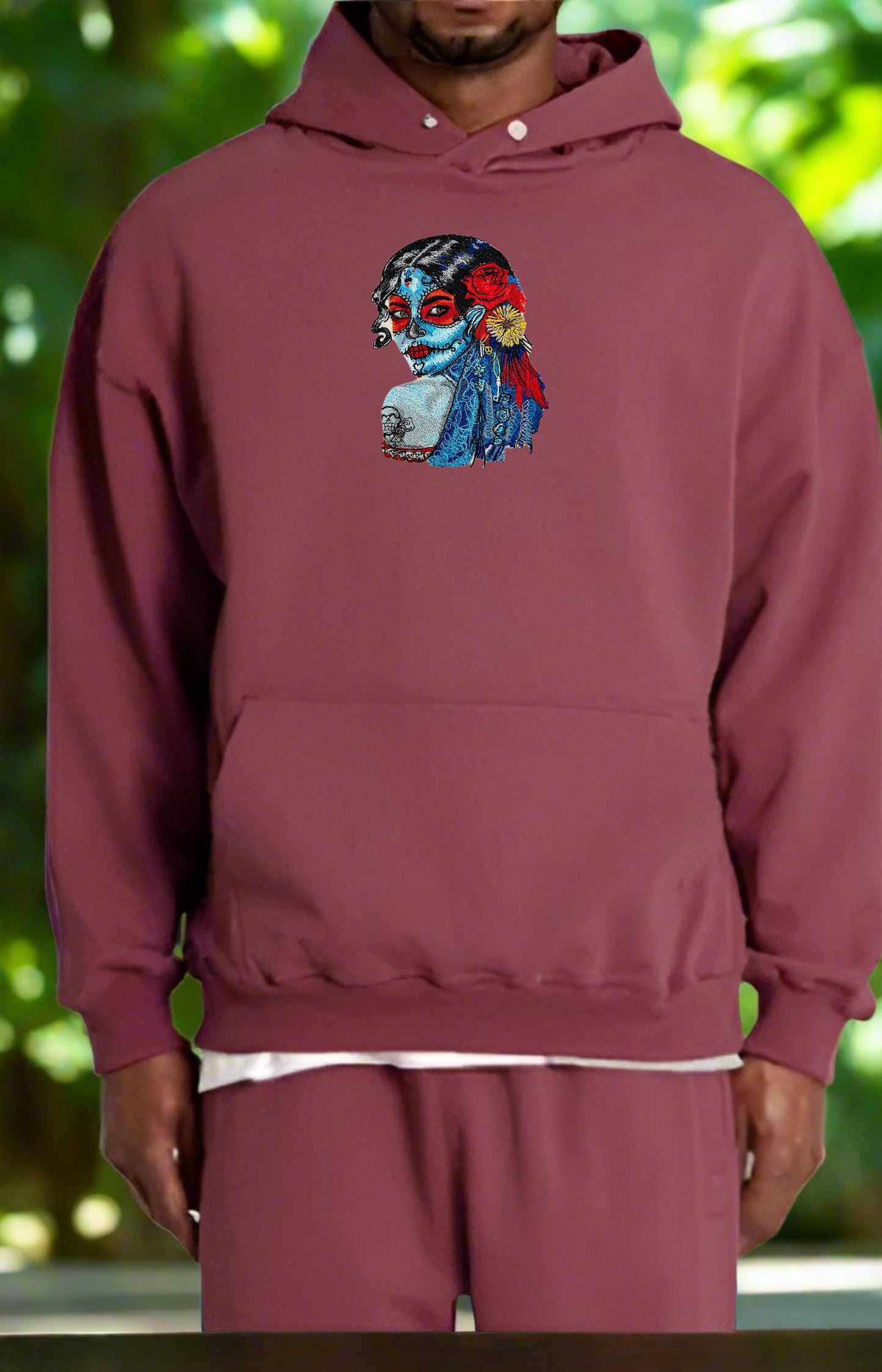 Maroon hoodie with a colorful Sugar Skull Lady Embroidery – Día de los Muertos Art design on a blurred green background