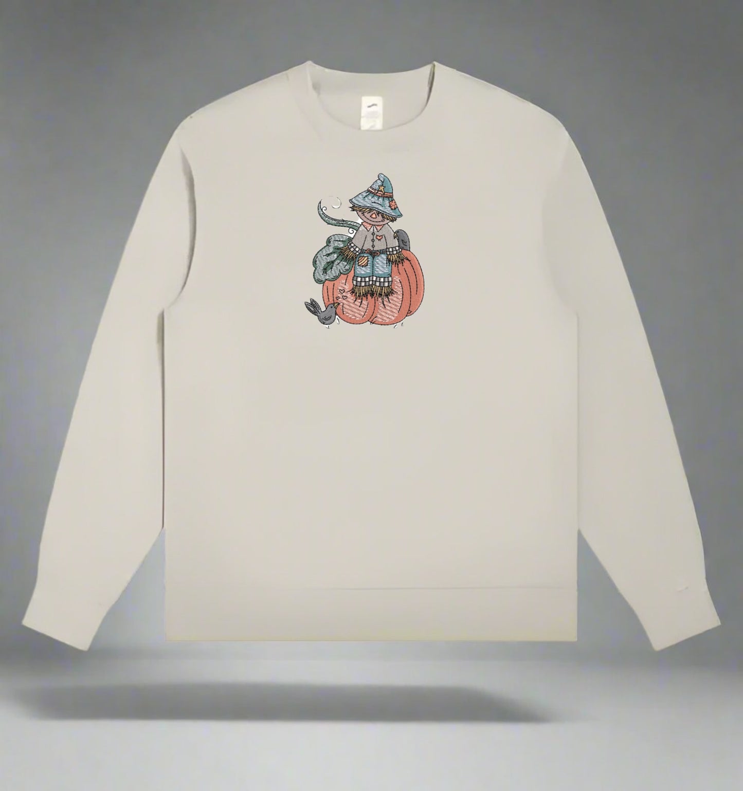 Beige long-sleeve shirt with a graphic of a person in a witch costume on a pumpkin against a gray background.