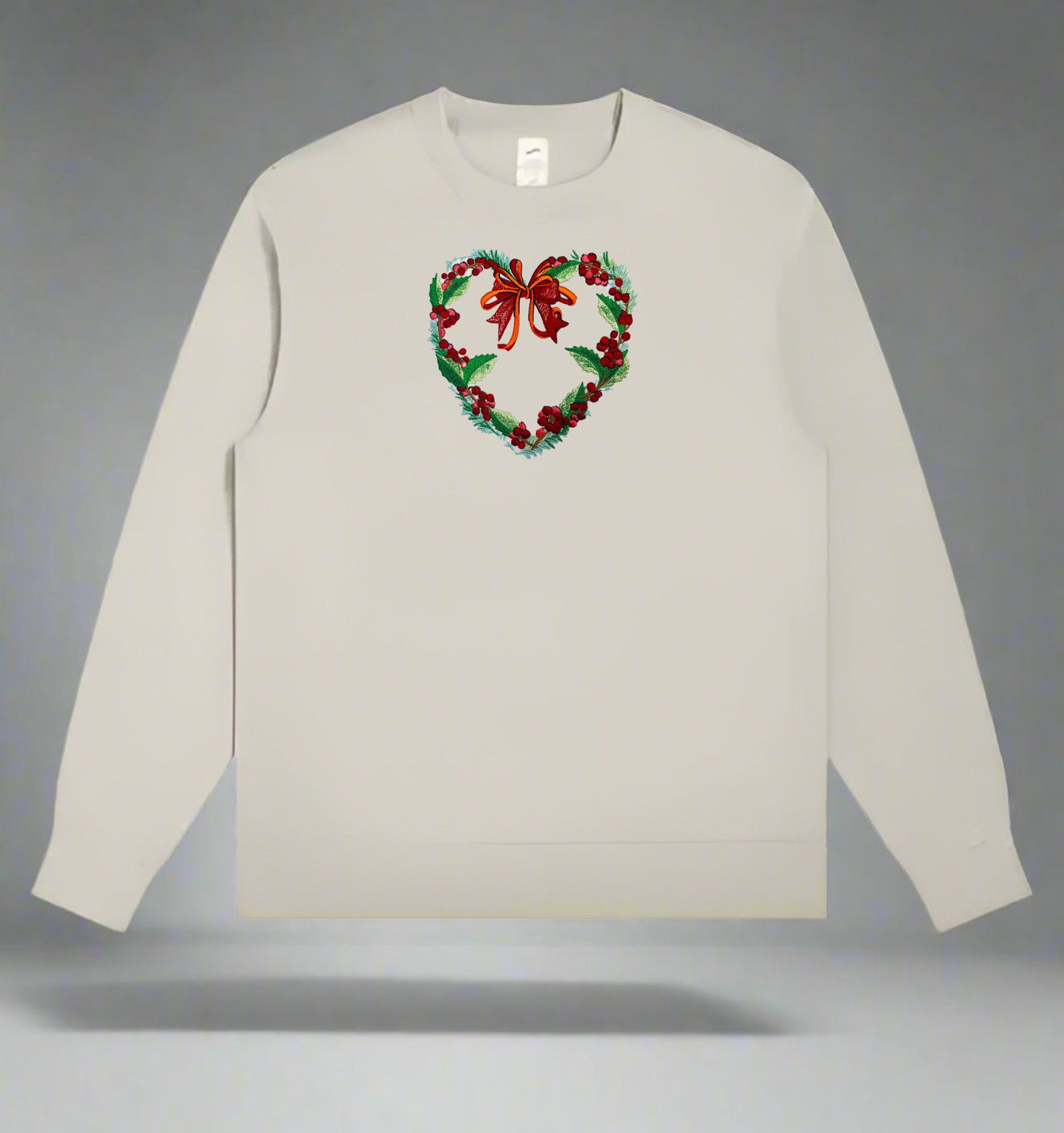 Light beige crewneck sweatshirt with an embroidered heart-shaped holiday wreath of green leaves, red berries, and a red bow centered on the chest.