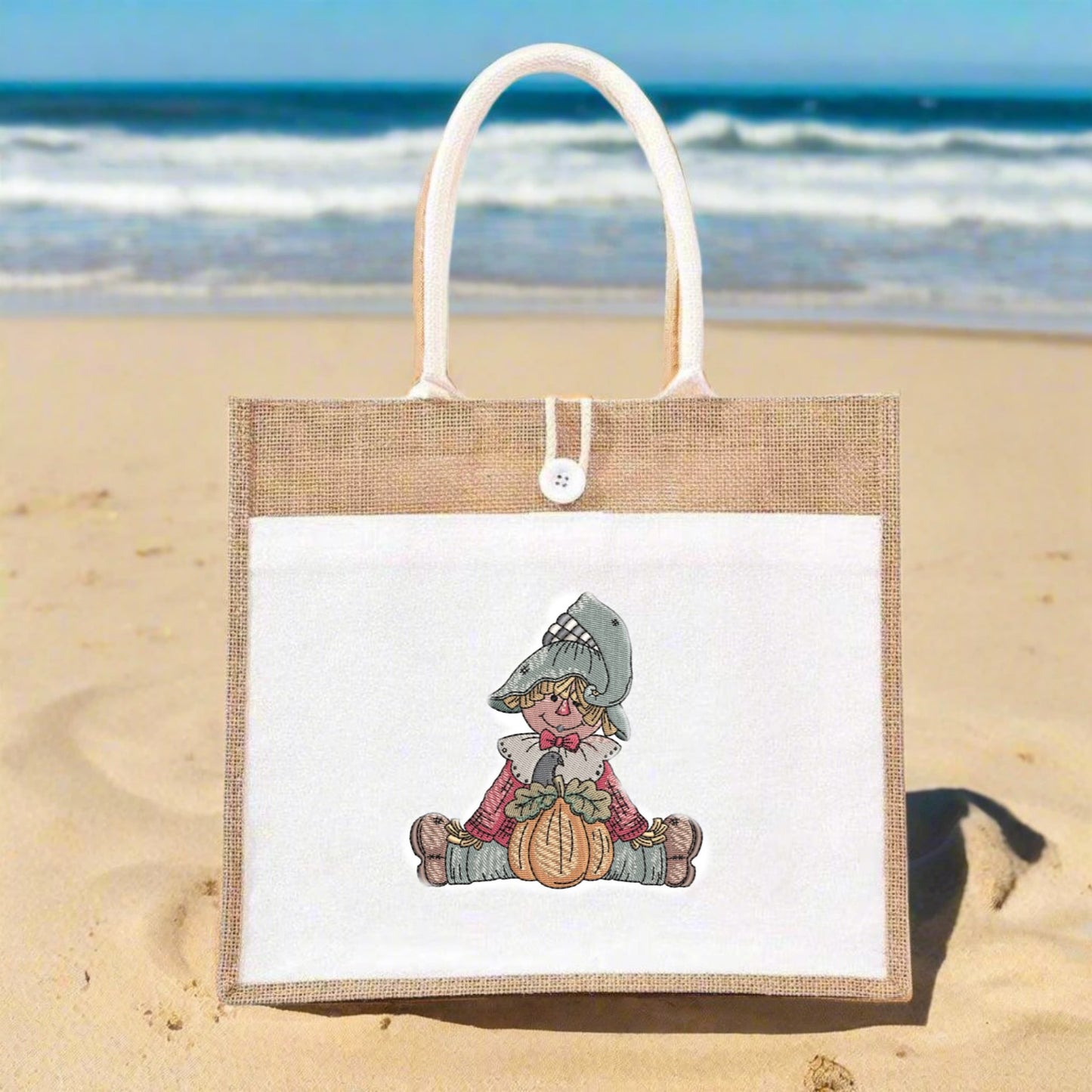Jute bag with a Scarecrow Pumpkin Embroidery Design on a sandy beach with ocean in the background
