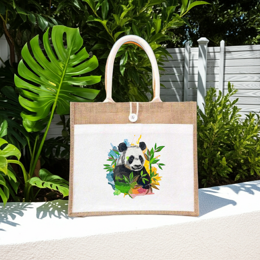 Natural jute tote handbag featuring a color splash panda embroidery design, displayed outdoors on a wooden bench with lush green tropical plants and a white slatted backdrop.