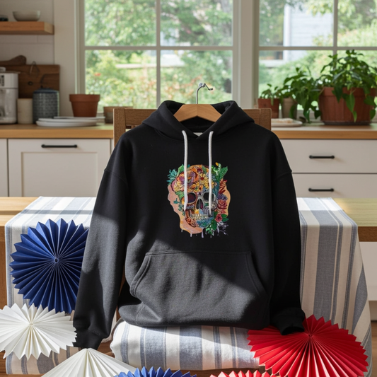 Black hoodie mockup featuring an embroidered colorful floral skull design on the chest, displayed on a chair in a bright kitchen with large windows, a striped tablecloth, and red, white, and blue paper fan decorations.