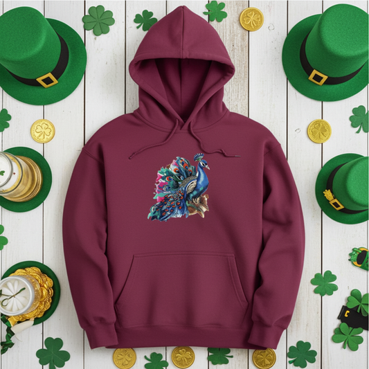 Burgundy hoodie mockup featuring an embroidered blue peacock with colorful feather details on the chest, styled flat lay on a white wood background with green shamrocks, green hats, and gold coins.