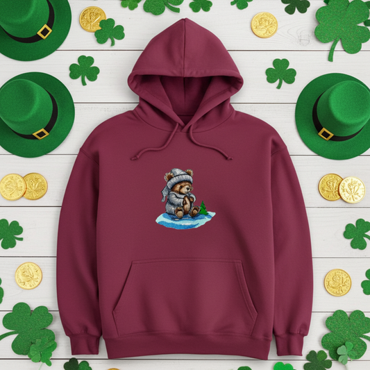 Burgundy premium hoodie with Moonlit Bear embroidery (teddy bear in winter hat and scarf on snowy ground), styled on a wooden background with green shamrock decorations, hats, and gold coins, original EmbroMania artwork.
