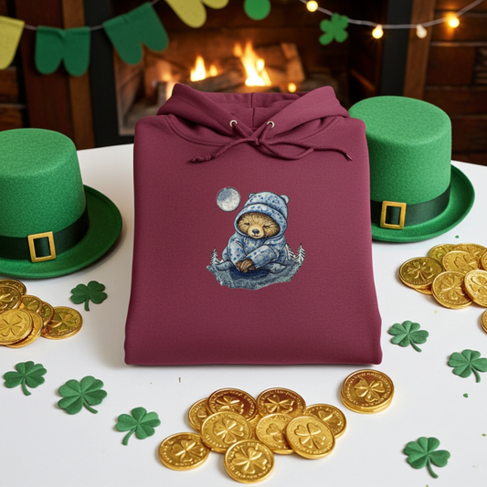Burgundy premium hoodie with Moonlit Sloth embroidery (sleepy sloth in a blue winter hoodie under a full moon), folded flat on a table with shamrock decor, green hats, gold coins, and a cozy fireplace background, original EmbroMania artwork.