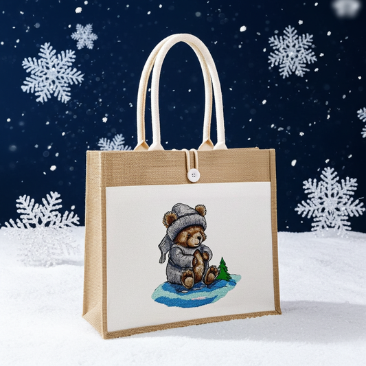 Natural jute tote handbag featuring a moonlit winter bear embroidery design, photographed in a snowy scene with falling snow and large snowflake decorations on a dark blue background.