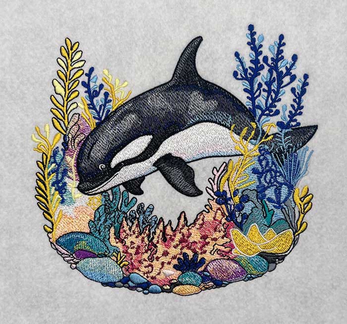 Embroidered design of an orca surrounded by colorful coral and seaweed on a light gray background.