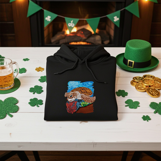 Black premium hoodie with Sea Turtle & Coral Reef embroidery showing a sea turtle above red coral and green sea plants, folded on a table with shamrock decorations, gold coins, and a green hat near a fireplace, original EmbroMania artwork.