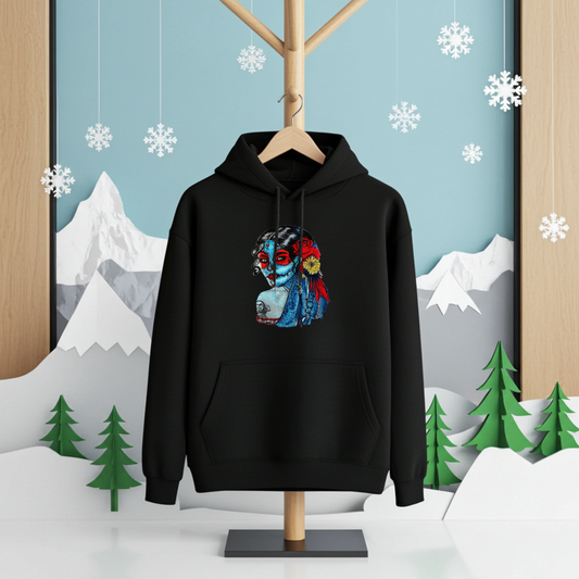 Black premium hoodie with Sugar Skull Lady embroidery featuring a Día de los Muertos portrait in blue and red tones with floral accents, hanging in a winter scene with snowflakes, mountains, and pine trees, original EmbroMania artwork.