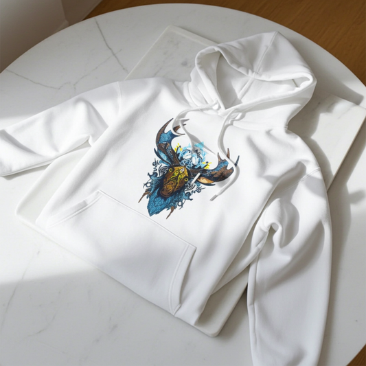 White premium hoodie with Tribal Moose embroidery featuring a moose head with bold antlers and tribal linework in deep blue, brown, and gold tones, styled flat-lay on a white marble table in natural sunlight, original EmbroMania artwork.