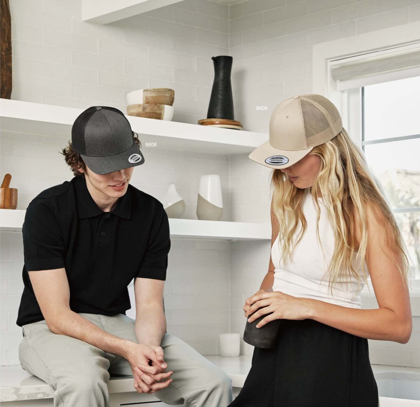 Two people wearing YP Classics trucker and snapback caps in a bright room; ideal base for custom 3D puff embroidery by EmbroMania, Dubai.