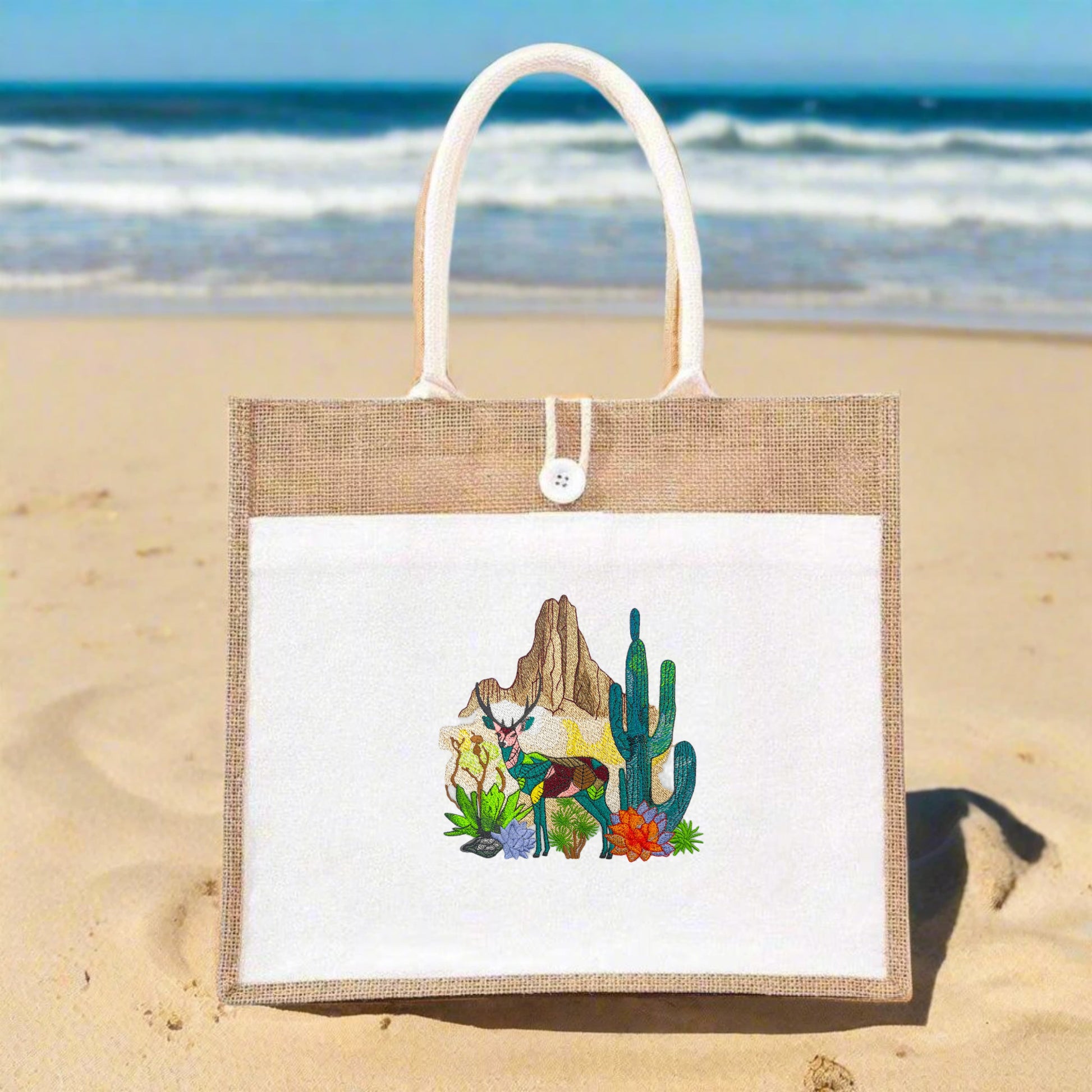 A jute tote bag with desert-themed embroidery including a deer, cacti, and flowers against a mountainous backdrop.