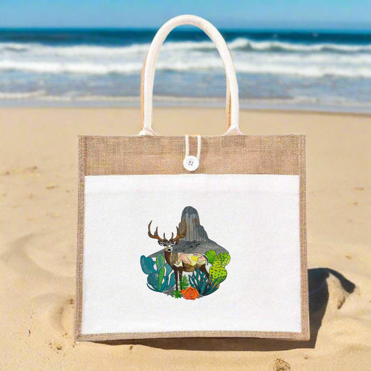 A jute tote bag with intricate embroidery of a mountain buck among floral and mountainous terrain.