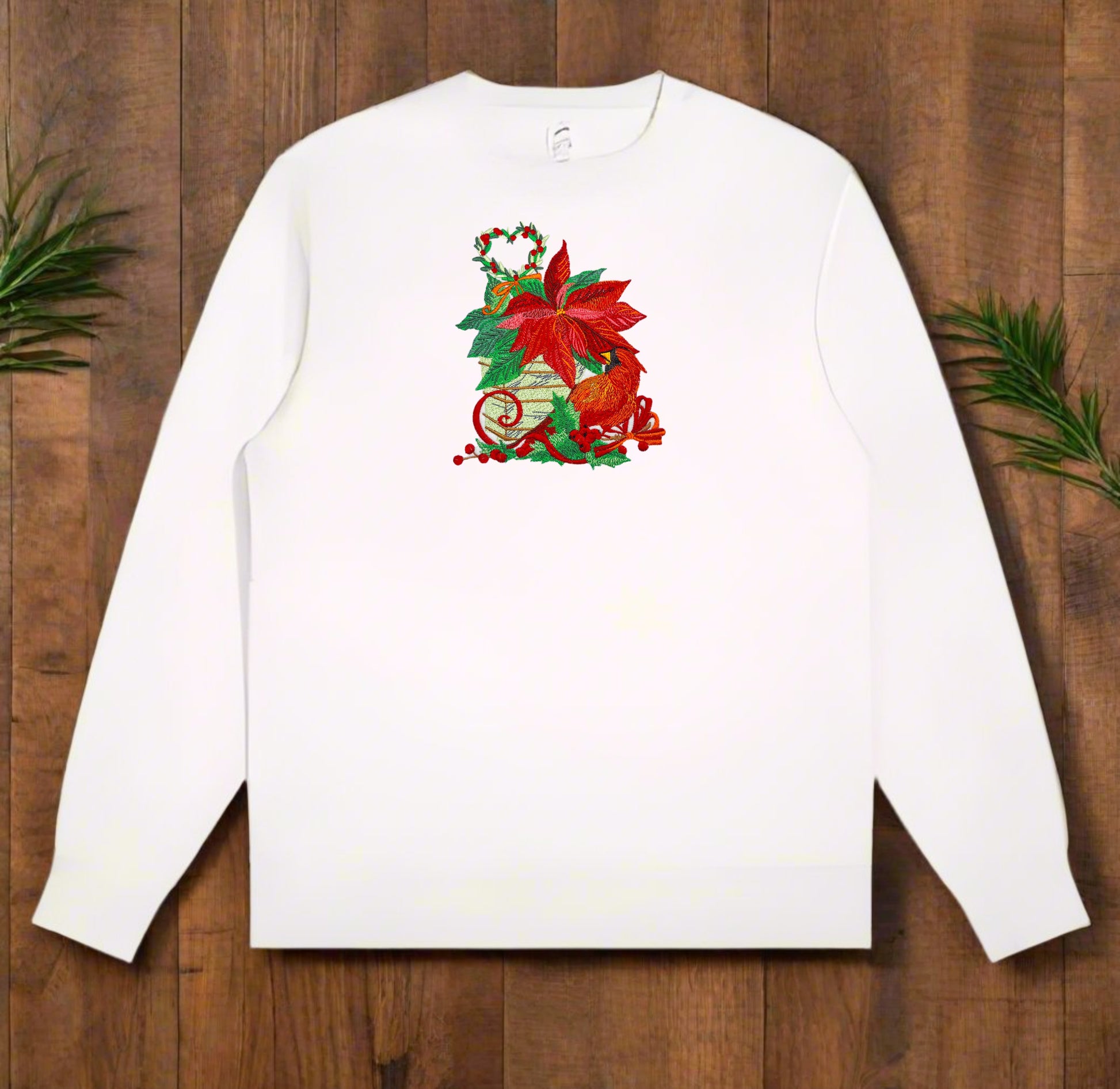 White crewneck sweatshirt on wood background with embroidered poinsettia bouquet, red cardinal, holly berries, pine foliage, and a heart-shaped mini wreath.