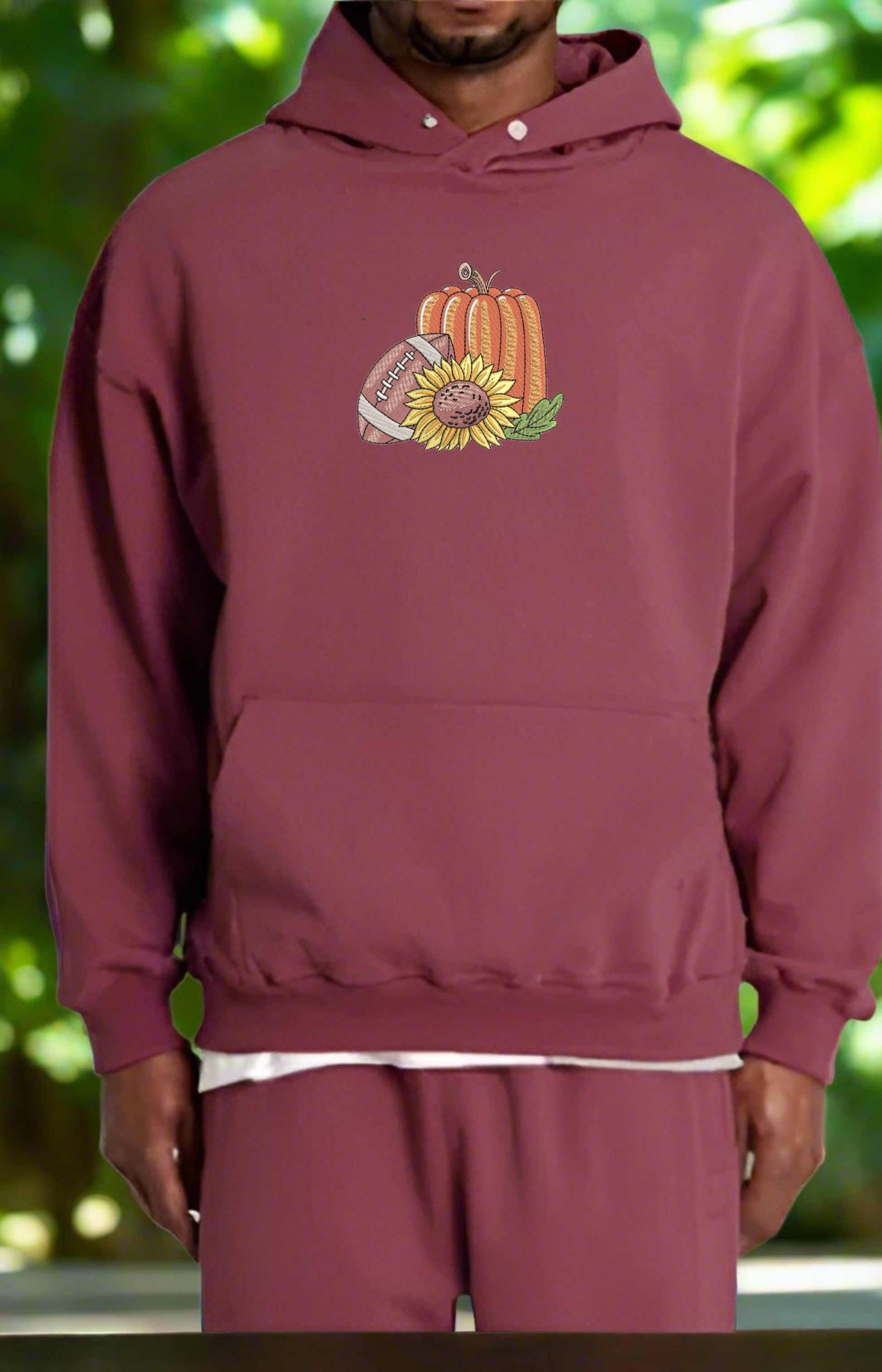 Maroon hoodie with a pumpkin and sunflower design worn by a person against a blurred green background
