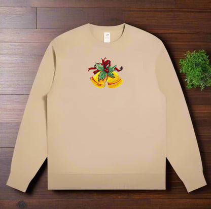 Tan crewneck sweatshirt laid on a wooden surface, featuring embroidered golden Christmas bells with green holly leaves and a red ribbon centered on the chest.