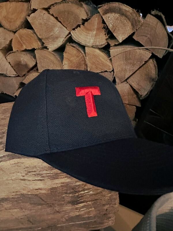 A black baseball cap with the letter 'T' embroidered in red, placed on a wooden surface with cut wood in the background.