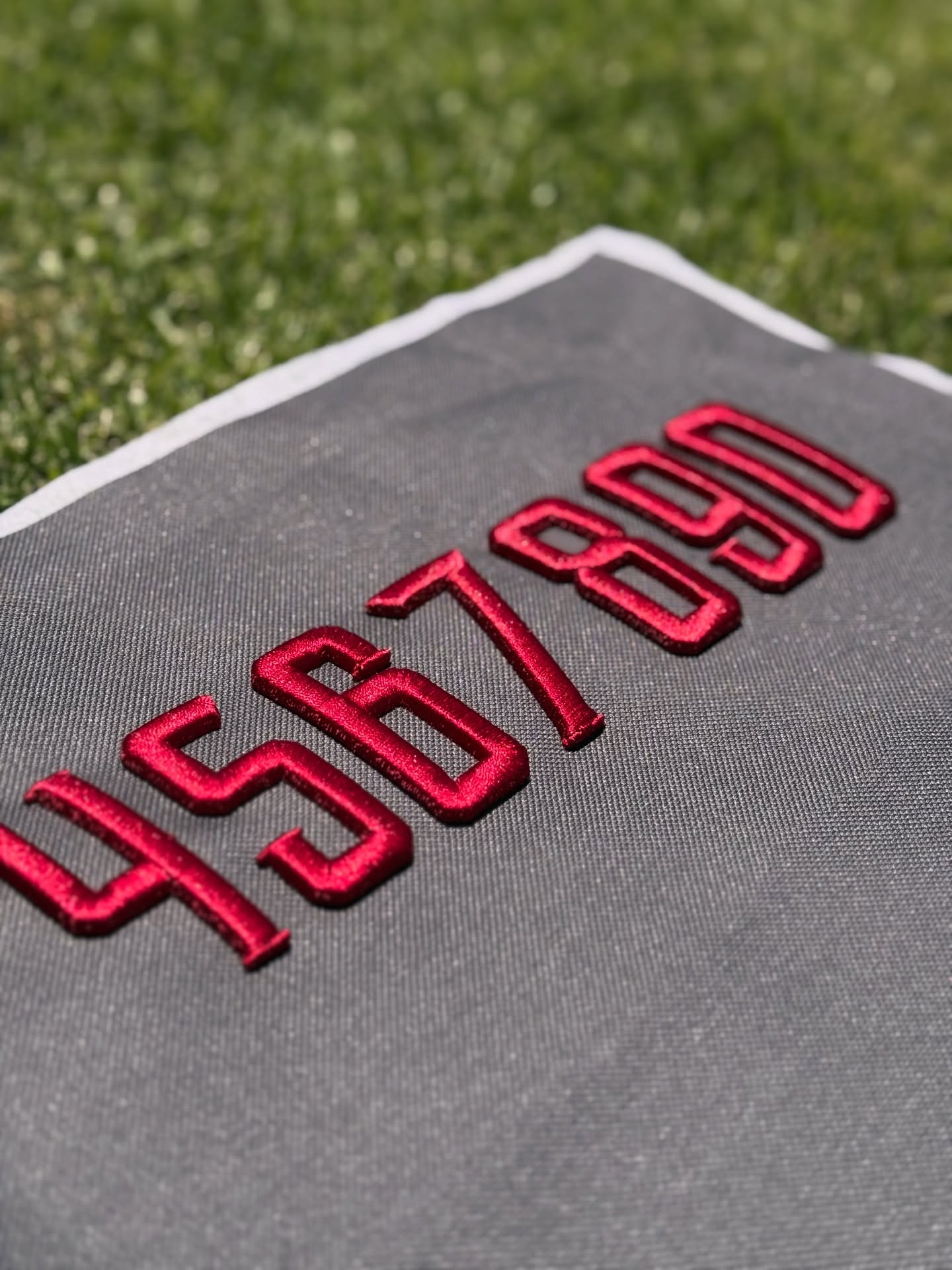 Close-up of red satin-stitch embroidered numbers “561890” in Metro Block lettering on dark gray fabric, photographed outdoors on grass.