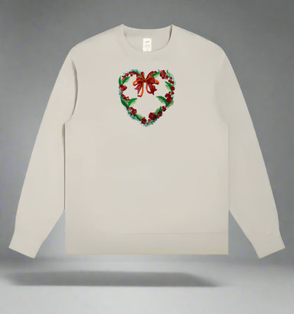 Light beige crewneck sweatshirt with an embroidered heart-shaped holiday wreath of green leaves, red berries, and a red bow centered on the chest.