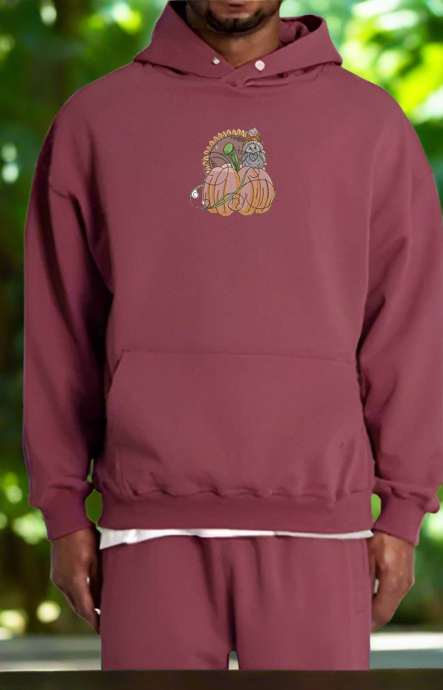 Maroon hoodie with a Pumpkin Bird & Sunflower Embroidery Design on a blurred green background