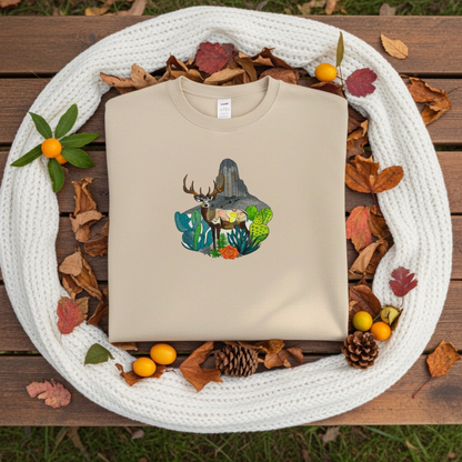 Soft beige sweatshirt folded on a wooden surface with a desert deer landscape embroidery design, styled with a white scarf, autumn leaves, pinecones, and yellow berries outdoors