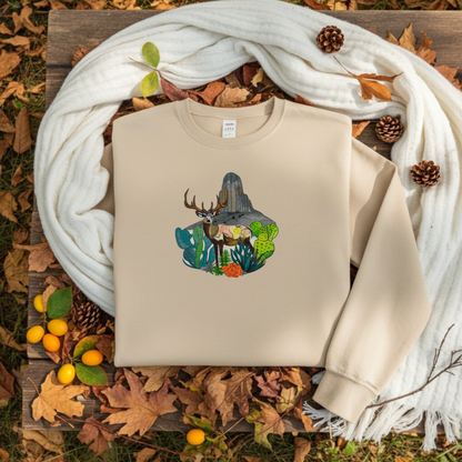 Soft beige sweatshirt folded on a wooden surface with a desert deer landscape embroidery design, styled with a white scarf, autumn leaves, pinecones, and yellow berries outdoors