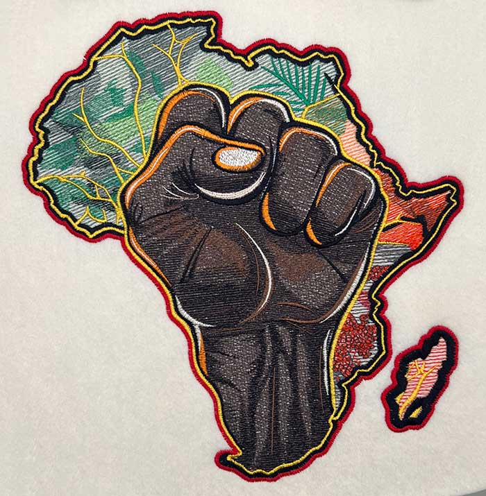 Close-up of an embroidered raised fist over a colorful Africa map outline, stitched on light fabric.