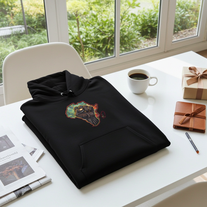 Black hoodie mockup featuring an embroidered raised fist over an Africa map design on the chest, folded on a white desk with coffee, notebook, pen, and gift box near a window.