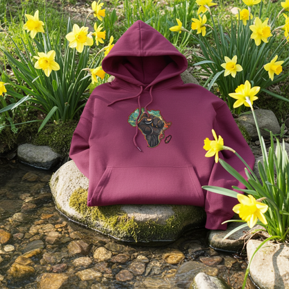 Burgundy hoodie mockup featuring an embroidered raised fist over an Africa map design on the chest, styled outdoors on a mossy rock in a shallow stream with yellow flowers.