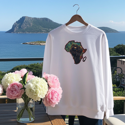 White sweatshirt featuring an Africa map with a raised unity fist embroidery-style design, displayed on a hanger on a balcony with ocean views and a vase of pink flowers.