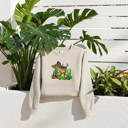 Mint green sweatshirt featuring an alpine desert fusion deer embroidery-style design with mountains, cactus, and colorful desert plants, displayed on a hanger outdoors with lush green leaves.