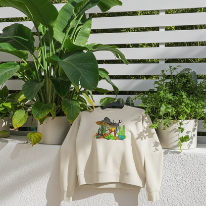 Mint green sweatshirt featuring an alpine desert fusion deer embroidery-style design with mountains, cactus, and colorful desert plants, displayed on a hanger outdoors with lush green leaves.