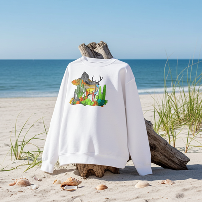 White sweatshirt featuring an alpine desert fusion deer embroidery-style design with mountains, cactus, and colorful desert plants, displayed on a hanger at the beach with seashells on a bench.