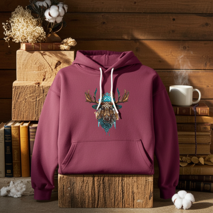 Burgundy hoodie mockup featuring an embroidered tribal-style moose head with turquoise accents on the chest, styled with books, cotton stems, and a warm rustic interior.