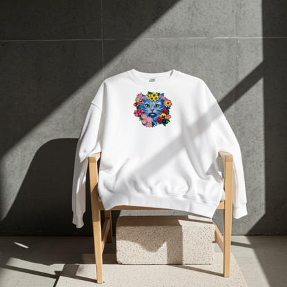 White sweatshirt draped on a wooden chair in a modern studio, featuring a blue cat face embroidery surrounded by colorful floral details on the chest.